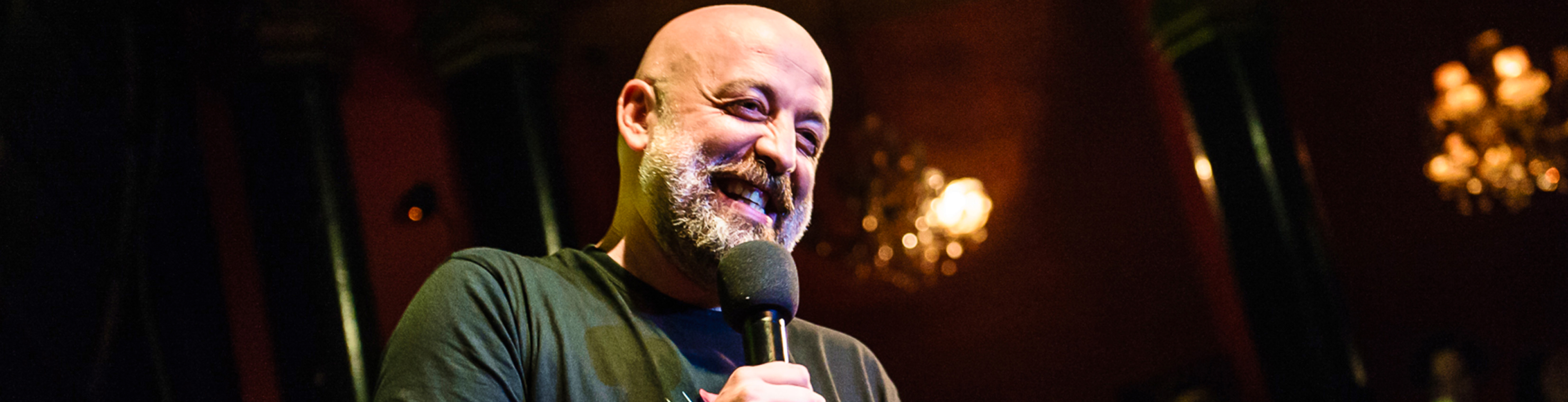 A smiling stand-up comedian holding a microphone on stage.